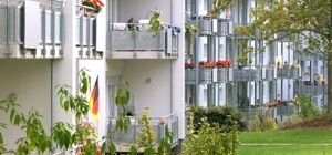 Links sanierte Gebäudezeile mit angebauten Balkonen, Block auf angelegte Grünanlage mit Bäumen und Sträuchern.