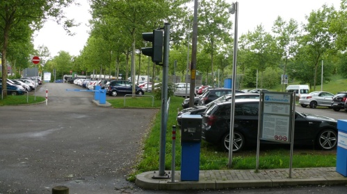 Einfahrt Parkplatz mit Gebührenhinweis, parkende Autos unter Bäumen, links Ausfahr mit Schranke.
