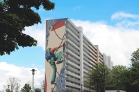 140 Meter langes Hochhaus mit 15 Etagen, saniert, vorne Laubengänge mit hellgrauen Brüstungen. Giebel mit Graffito, das einen Bergsteier zeigt.