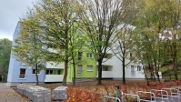 Viergeschossiges Wohnhaus mit Flachdach, Fassade blau, weiß und grün. Im Vordergrund Bäume im Frühling.