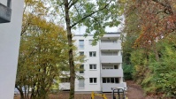 Wohnahus mit vier Etagen, Flachdach, Balkone, in weiß. Hohe Bäume davor. 