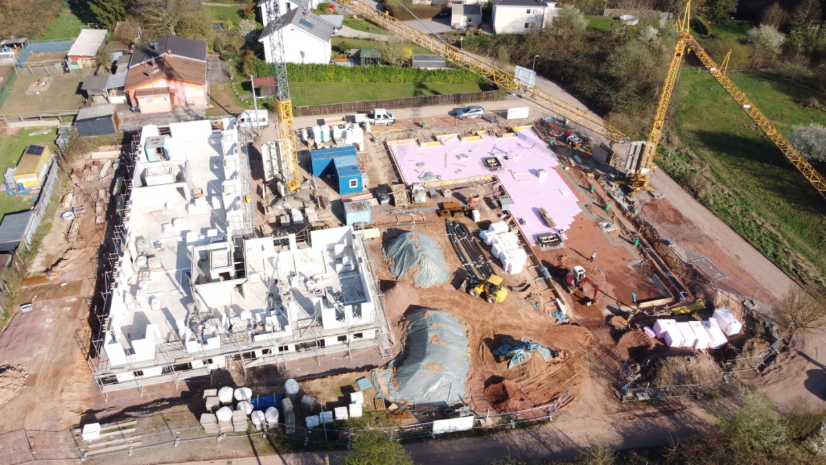 Drohnenaufnahme der Baustelle mit 1. Gebäude links mit Erdgeschoss und Mauern 1. Etage, hinten rechts Bodenplatte 2. Gebäude.